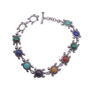 May include: Silver bracelet with turtle-shaped links and colourful gemstone accents. The bracelet has a toggle clasp and is made up of multiple turtle charms, each holding a different coloured stone, including green, blue, and red.