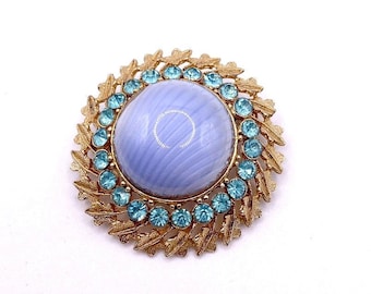 May include: A vintage brooch with a large, light blue center stone surrounded by small, round, turquoise-colored stones. The brooch has a gold-toned metal frame with a leaf design around the edge. A classic piece of jewelry.