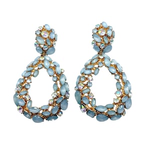 Peut inclure: Une paire de boucles d'oreilles pendantes dorées avec un design en forme de goutte ouverte, incrustées de pierres ovales bleu clair et de petits cristaux irisés. Les boucles présentent un amas de pierres bleues au niveau du fermoir.