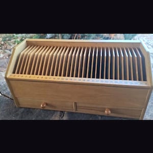 Puede incluir: Un organizador de escritorio de madera con múltiples ranuras verticales para almacenamiento. El organizador tiene dos cajones pequeños con pomos de madera y una escala numerada a lo largo del borde superior, del 1 al 31. La madera tiene un acabado marrón claro natural.