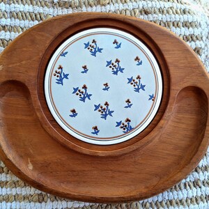 Puede incluir: Bandeja ovalada de madera con un inserto de cerámica blanca. El inserto presenta un estampado floral con flores azules y marrones. La bandeja tiene dos hendiduras curvas. La madera tiene un acabado cálido y natural.