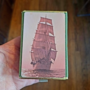 Könnte beinhalten: Eine rechteckige Schachtel mit grünem Rand und goldener Zierleiste. Die Schachtel zeigt ein Vintage-Bild eines Großseglers, der auf dem Meer segelt. Das Schiff hat mehrere Segel und ist vor einem rosafarbenen Himmel abgebildet.