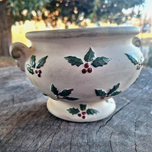 Puede incluir: Cuenco de cerámica color crema con base de pedestal y dos asas laterales. El cuenco está decorado con hojas verdes de acebo y bayas rojas. El borde y las asas tienen un ribete gris-marrón. El cuenco está sobre una superficie de madera.