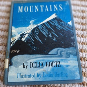 May include: A vintage book titled "Mountains" by Delia Goetz, illustrated by Louis Darling. The cover features a blue sky with a snow-capped mountain illustration. The book has a blue border and a library sticker.