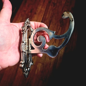 May include: Ornate silver-colored wall hook with a decorative backplate and a curved hook design. The hook features intricate detailing and a vintage aesthetic, suitable for hanging items. The hook is made of metal.