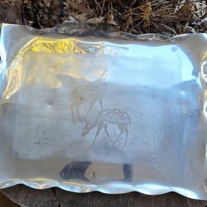 May include: A rectangular, silver-toned metal serving tray with a wavy, scalloped edge. The tray features an etched design of two deer in a field. The deer are depicted in a naturalistic style, with one grazing and the other standing.