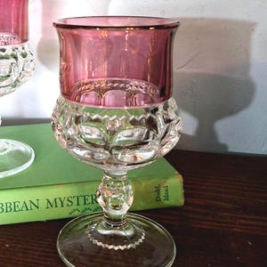 May include: A clear glass goblet with a pink rim and bowl. The bowl has a cut glass design, and the stem is clear with a round base. The goblet is on a wooden surface. A book titled "Caribbean Mystery" is in the background.