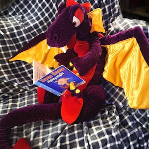 Pode incluir: Um brinquedo de pelúcia de dragão roxo com detalhes vermelhos e asas amarelas, lendo um livro intitulado "Llama Llama Nighty-Night". O dragão está sentado em um cobertor xadrez.