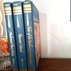 Puede incluir: Tres libros antiguos de tapa dura con cubiertas azules y letras doradas. Los títulos incluyen "Andersen's Fairy Tales", "Treasure Island" y "Tales of the Brothers Grimm". Los libros se exhiben sobre una superficie de madera.