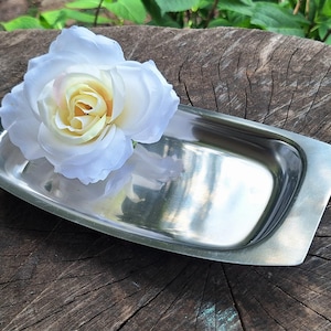 May include: A rectangular stainless steel tray with a polished, reflective surface and slightly tapered ends. A single large, artificial white rose with a pale yellow center rests inside the tray, partially covering the metallic surface.
