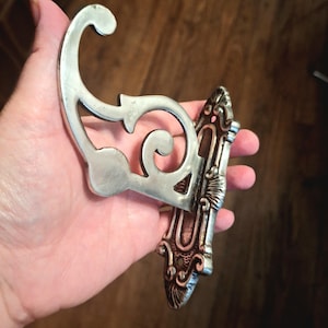 Könnte beinhalten: Verzierter silberfarbener Wandhaken mit dekorativem Scroll-Design. Der Haken hat eine geschwungene Oberseite und eine detaillierte Rückplatte mit floralen Akzenten. Ein Einrichtungsgegenstand im Vintage-Stil.