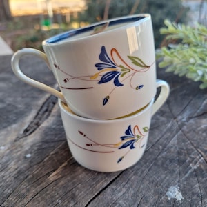 Puede incluir: Dos tazas de té de cerámica blanca apiladas con diseños florales en azul, naranja y verde. Las tazas tienen un borde azul oscuro y un asa curva. Las tazas están apiladas sobre una superficie de madera desgastada.