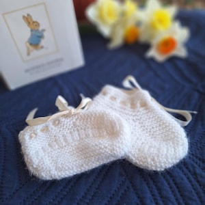 May include: A pair of white knitted baby mittens with cream-colored ribbon ties. The mittens are on a navy blue textured blanket, with a copy of "The Tale of Peter Rabbit" and daffodils in the background.