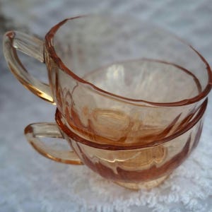 Könnte beinhalten: Zwei übereinander gestapelte Teetassen aus bernsteinfarbenem Glas mit geriffeltem Design. Die Tassen haben eine abgerundete Form und einen Henkel.