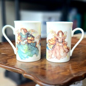 May include: Two white ceramic mugs featuring watercolour-style illustrations of winged figures. The left mug displays a figure in a flowing teal gown, while the right mug shows a figure in a flowing mauve gown, both holding small golden instruments.