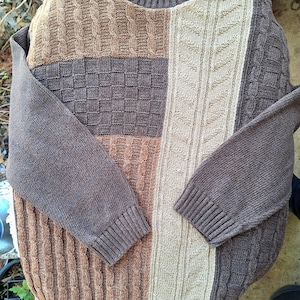 May include: A patchwork-style sweater in shades of brown, tan, and gray. The sweater features various knit patterns, including cable knit and a checkered design. The sleeves and collar are a solid gray color. The label reads "Liberty Sweaters".