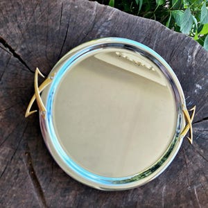 May include: A circular, silver-coloured serving tray with gold-coloured handles. The tray has a reflective surface and a blue accent around the rim. The handles are shaped in a decorative, angular design.