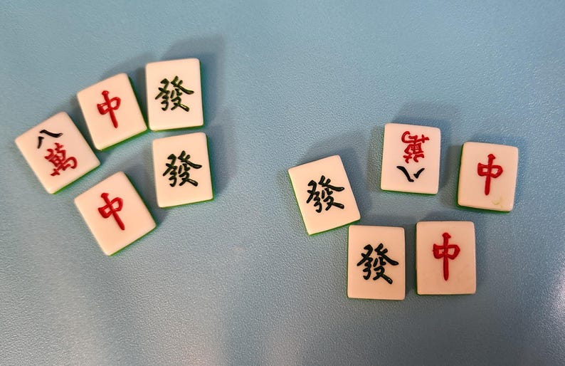 K&ouml;nnte beinhalten: Elfenbeinfarbene Mahjong-Steine mit schwarzen und roten chinesischen Schriftzeichen liegen auf einer hellblauen Oberfl&auml;che. Die Steine zeigen Schriftzeichen wie "中", "發" und "八萬".