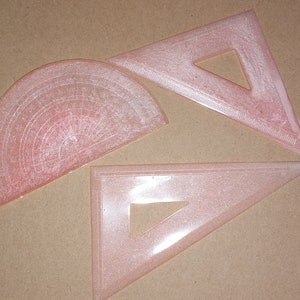 May include: Three pink plastic protractors, one is a half circle and the other two are triangles.