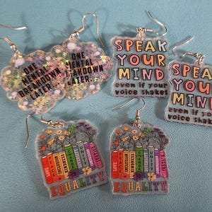 May include: A collection of five pairs of acrylic earrings. Two pairs feature the text "Speak Your Mind" and "One Mental Breakdown Later." The other two pairs have the word "Equality" and colorful books. The earrings have silver hooks.