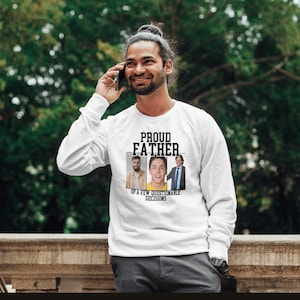 Op de afbeelding: Wit sweatshirt met de tekst "PROUD FATHER" boven drie ingelijste foto's en de tekst "OF A FEW QUESTIONABLE DECISIONS". Het sweatshirt wordt door een persoon gedragen.