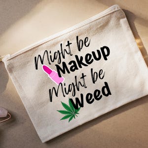 May include: A beige canvas makeup bag with a zip. The bag has the text "Might be Makeup Might be Weed" in black, a pink lipstick graphic, and a green cannabis leaf.