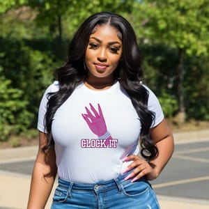 May include: White t-shirt with a pink hand graphic and the text "CLOCK IT". The hand is adorned with a bracelet. The model is wearing blue jeans.