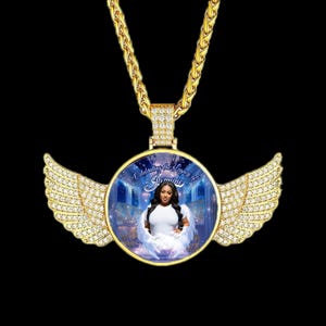 Puede incluir: Collar con colgante dorado con un dije redondo y alas con incrustaciones de diamantes. El dije muestra una foto de una mujer con un vestido blanco y el texto "Celebrating the Legacy of Jasmine". La cadena es gruesa y texturizada.