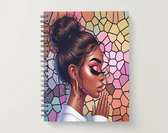Op de afbeelding: Spiraalgebonden notitieboek met een illustratie van een vrouw met donker haar in een knot, gouden oorringen en biddende handen. De achtergrond is een glas-in-loodmozaïek in roze, oranje en geel.