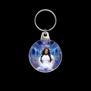 Puede incluir: Un llavero redondo plateado con un colgante circular. El colgante muestra a una mujer con alas de &aacute;ngel sobre un fondo azul. El texto dice "Celebrating the Legacy of Jasmine". La mujer lleva una camiseta blanca.