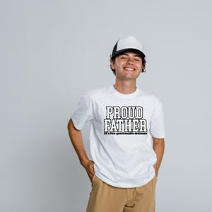 Op de afbeelding: Wit T-shirt met de tekst "PROUD FATHER" in een vetgedrukte, omlijnde letter. Daaronder staat de zin "Of a few questionable decisions". De persoon draagt een zwart-witte truckerpet en een kaki broek.