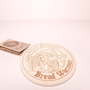 May include: Off-white ceramic bread warmer with a brown illustration of a cornucopia, grapes, and other fruits. The words "Natures Cornucopia" and "Bread Warmer" are printed on the warmer. A tag with the words "Bread & Bun Warmers" is also present.