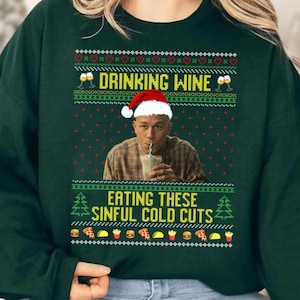 May include: Dark green sweatshirt with a festive design. The text "DRINKING WINE" and "EATING THESE SINFUL COLD CUTS" is displayed above and below a man wearing a Santa hat. The design includes images of wine glasses, food, and Christmas trees.