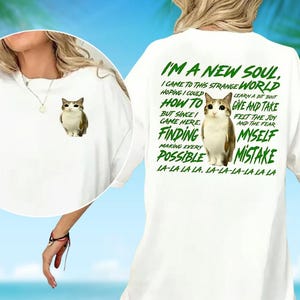 May include: White t-shirt featuring a cat graphic on the front and back. The back of the shirt has green text that reads "I'M A NEW SOUL" and other phrases. The shirt is a casual, oversized style.