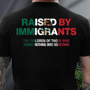 May include: Black t-shirt with the text "RAISED BY IMMIGRANTS" in green, white, and red. The shirt also features the phrase "THE CHILDREN OF THOSE WHO TURNED NOTHING INTO SOMETHING" in white and red.