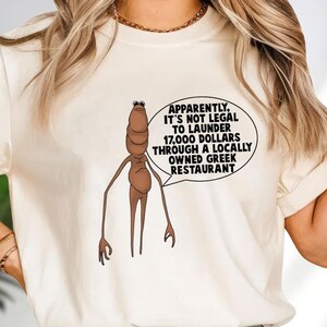 May include: Cream-colored t-shirt featuring a cartoonish brown figure with a speech bubble. The speech bubble contains the text: "Apparently, it's not legal to launder 17,000 dollars through a locally owned Greek restaurant."