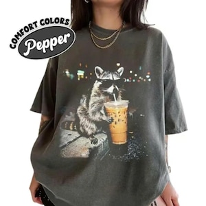 May include: Dark grey t-shirt with a graphic of a raccoon wearing sunglasses and drinking iced coffee. The shirt has the words "Comfort Colors Pepper" in a white oval on the upper left.