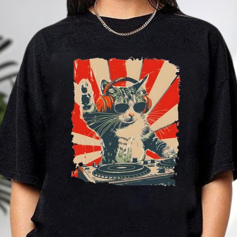 A black t-shirt with a graphic design depicting a cat wearing headphones and playing a turntable, with a red and white sunburst background.