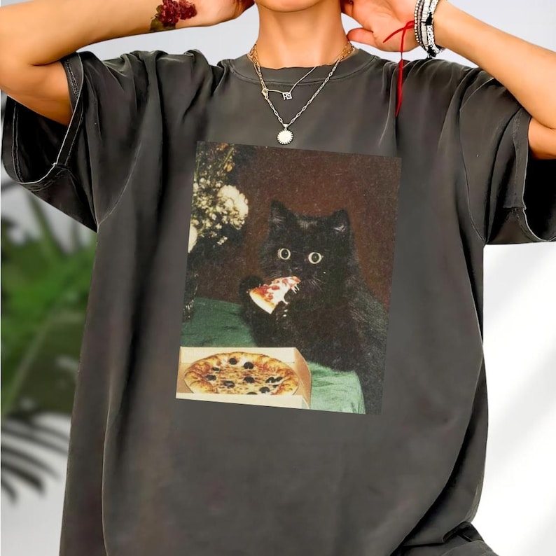 A person wearing a gray t-shirt with a printed image of a black cat eating pizza.