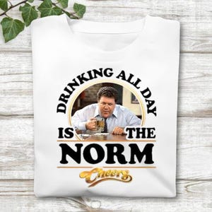 May include: White t-shirt featuring a graphic of a man holding a beer mug, with the text "DRINKING ALL DAY IS THE NORM" in black. Below the text is the word "Cheers" in gold script. The shirt is folded.