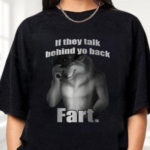 May include: Black t-shirt with a graphic of a wolf holding a hat, with the text "If they talk behind yo back Fart." The wolf is gray and white, with a smug expression. The text is in a bold, sans-serif font.