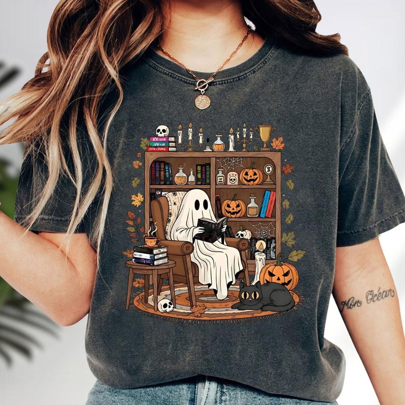 A woman wearing a t-shirt with a graphic design depicting a ghost reading a book in a library surrounded by Halloween-themed elements such as pumpkins, bats, and candles.