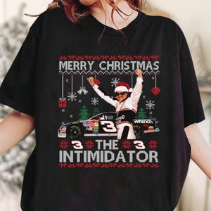 May include: Black Christmas t-shirt with the text "MERRY CHRISTMAS" and "3 THE 3 INTIMIDATOR". The shirt features a race car and a person wearing a Santa hat. Festive design with snowflakes, hearts, and Christmas trees.