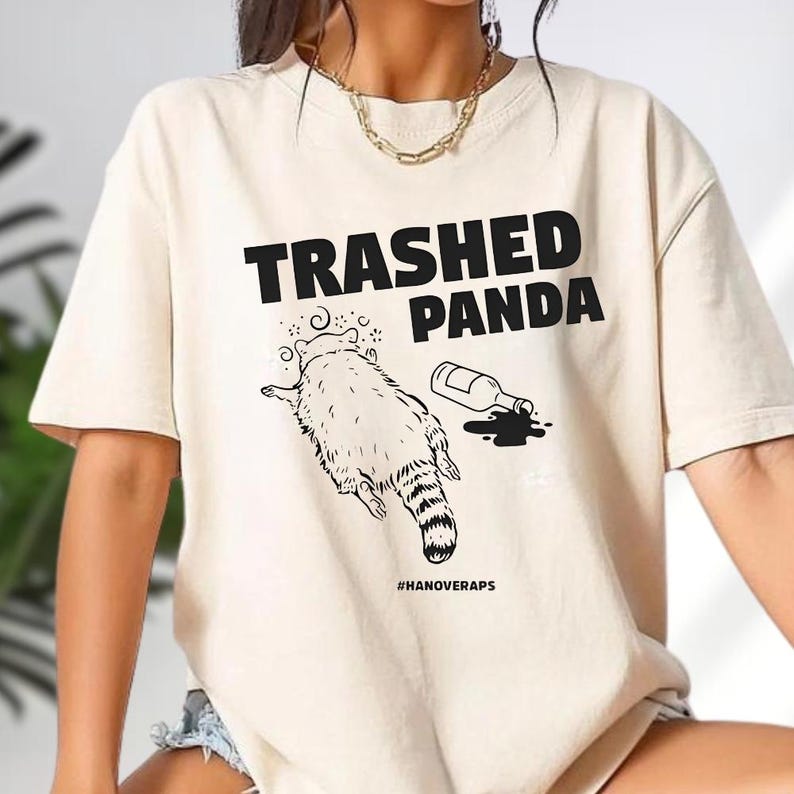 A person wearing a white t-shirt with a graphic design featuring a raccoon and text that says "Trashed Panda".