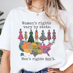 May include: A white short-sleeved t-shirt featuring a graphic of a multicolored map of the United States. Above the map, the text reads "Women's rights vary by state." Below the map, the text reads "Men's rights don't." Seven figures in dresses stand on the map.