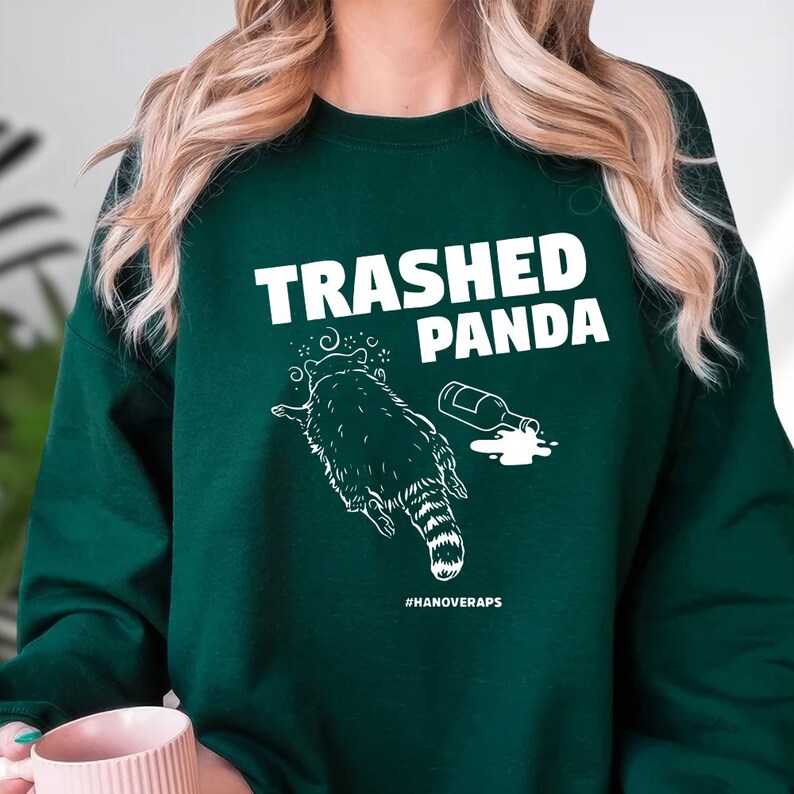 A person wearing a green sweatshirt with a graphic design that says "Trashed Panda" and features a raccoon.