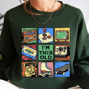 May include: Dark green sweatshirt with a retro-themed graphic. The graphic features nine squares with images of a boombox, joystick, TV, cassette tape, Polaroid camera, telephone, roller skate, and skateboard. The text "I'M THIS OLD" is in the center.