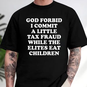 May include: A black short-sleeved t-shirt featuring white text printed on the chest that reads: GOD FORBID I COMMIT A LITTLE TAX FRAUD WHILE THE ELITES EAT CHILDREN. The shirt has a crew neckline and a standard fit, displaying bold, capitalized lettering.