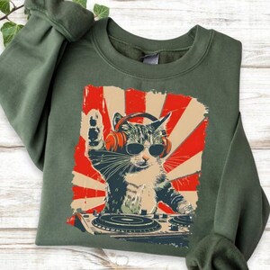 A green sweatshirt with a graphic design depicting a cat wearing sunglasses and headphones, appearing to be playing a musical instrument, such as a drum or guitar.