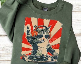 May include: Green sweatshirt featuring a graphic of a cat wearing sunglasses and headphones, operating a DJ mixer. The cat is set against a red and tan burst design. The sweatshirt is made of a soft material.